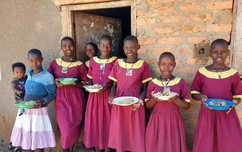 Girls in school uniforms holding bowls of food, one holding a baby